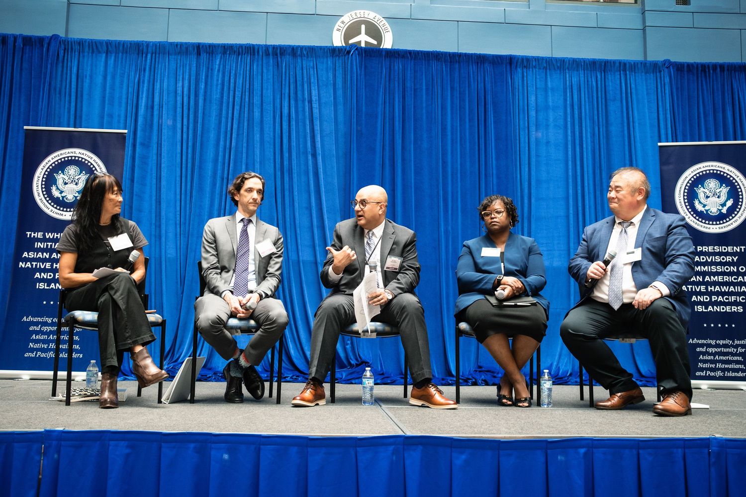 Five panelists seated on stage at a formal conference with blue curtains and presidential banners in background.