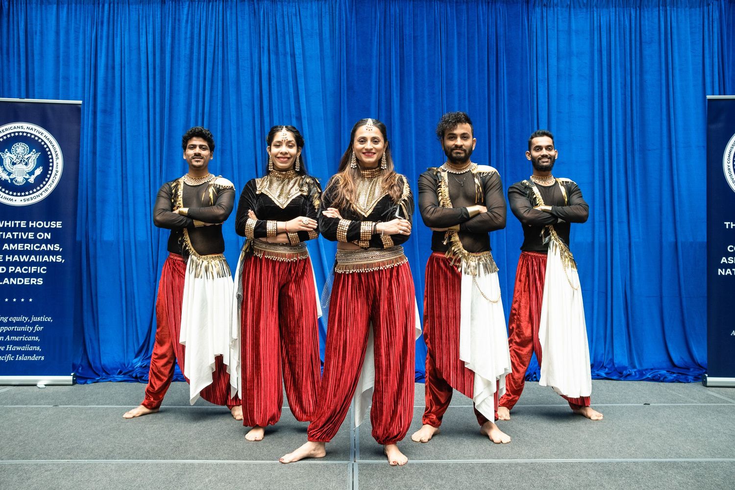 Five dancers in red and gold costumes pose on stage at a White House Initiative on Americans event.
