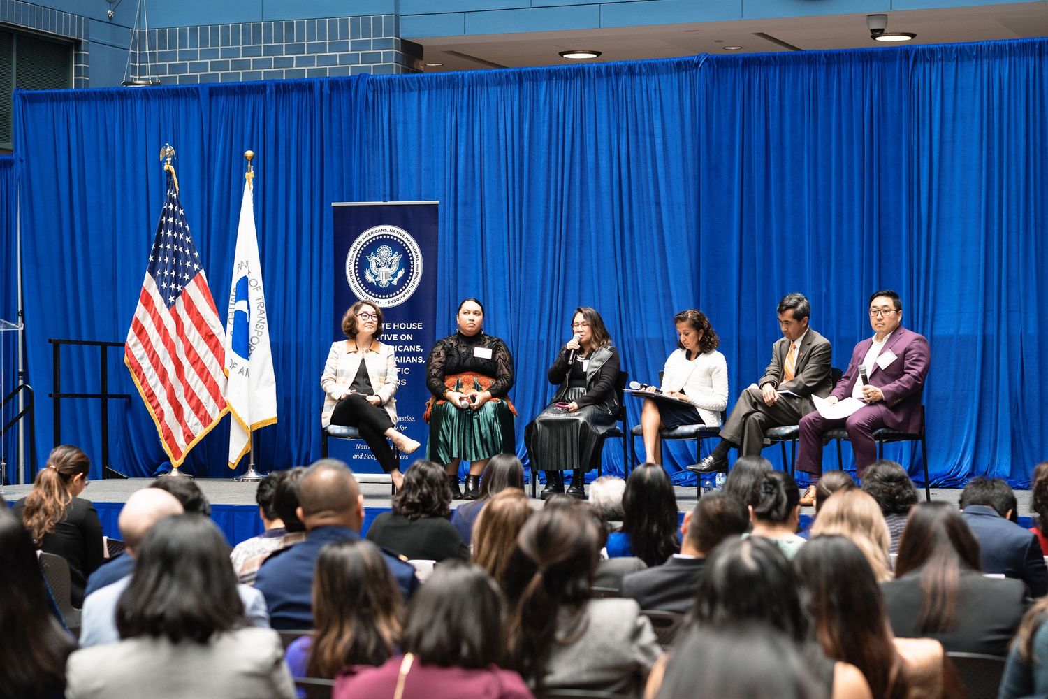 Panel discussion on stage with six speakers before a large audience at a formal government event with US flag.