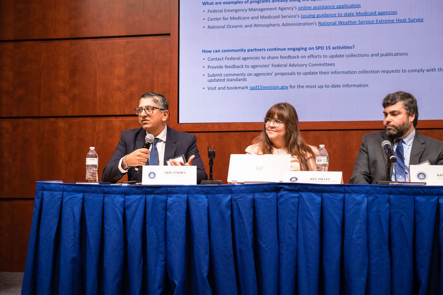 Three professionals sit at a panel discussion table with microphones, presenting in front of a informational display board.