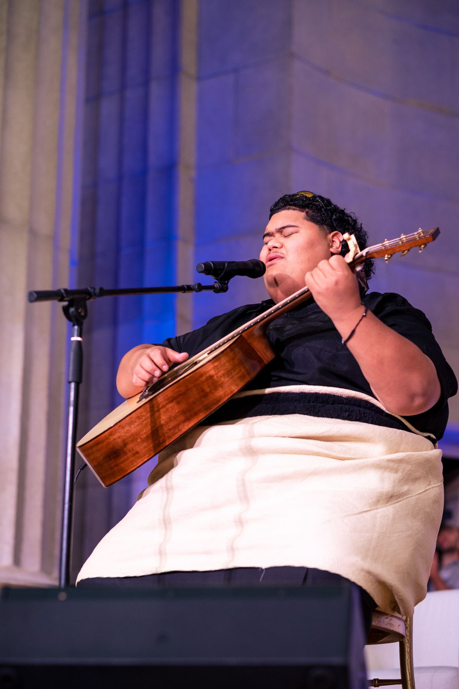 Young musician plays acoustic guitar and sings into microphone on stage, wearing black shirt and white wrap skirt.