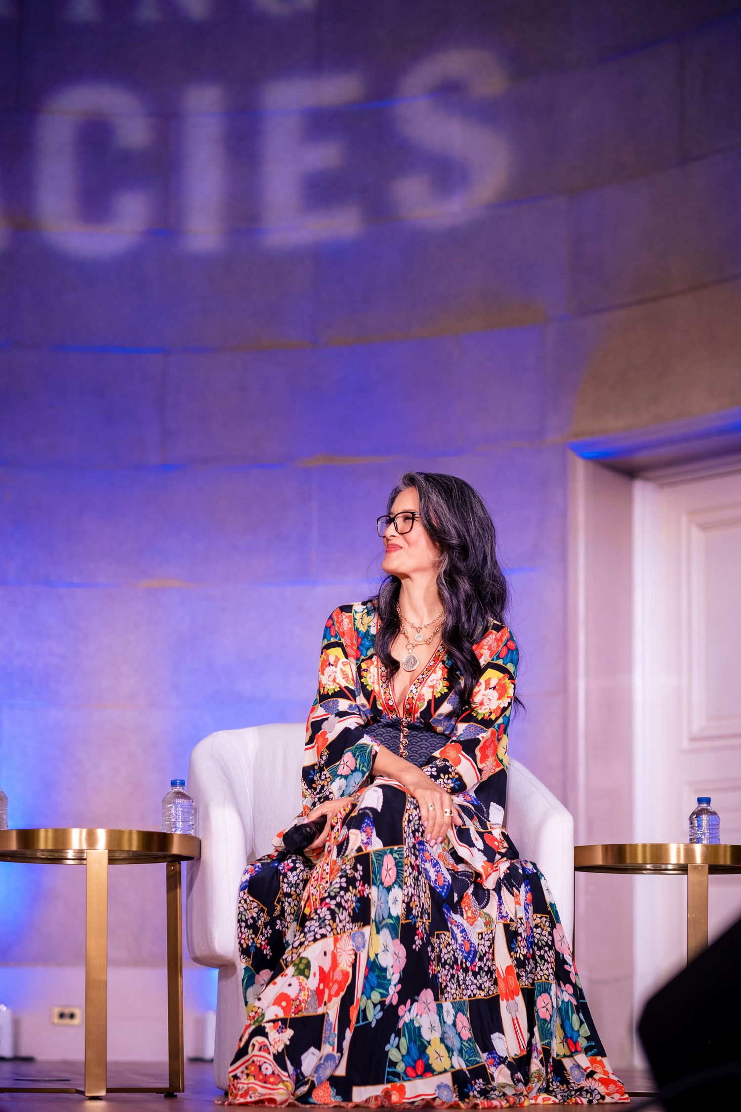 Woman in floral maxi dress seated on white chair on stage at a conference event.