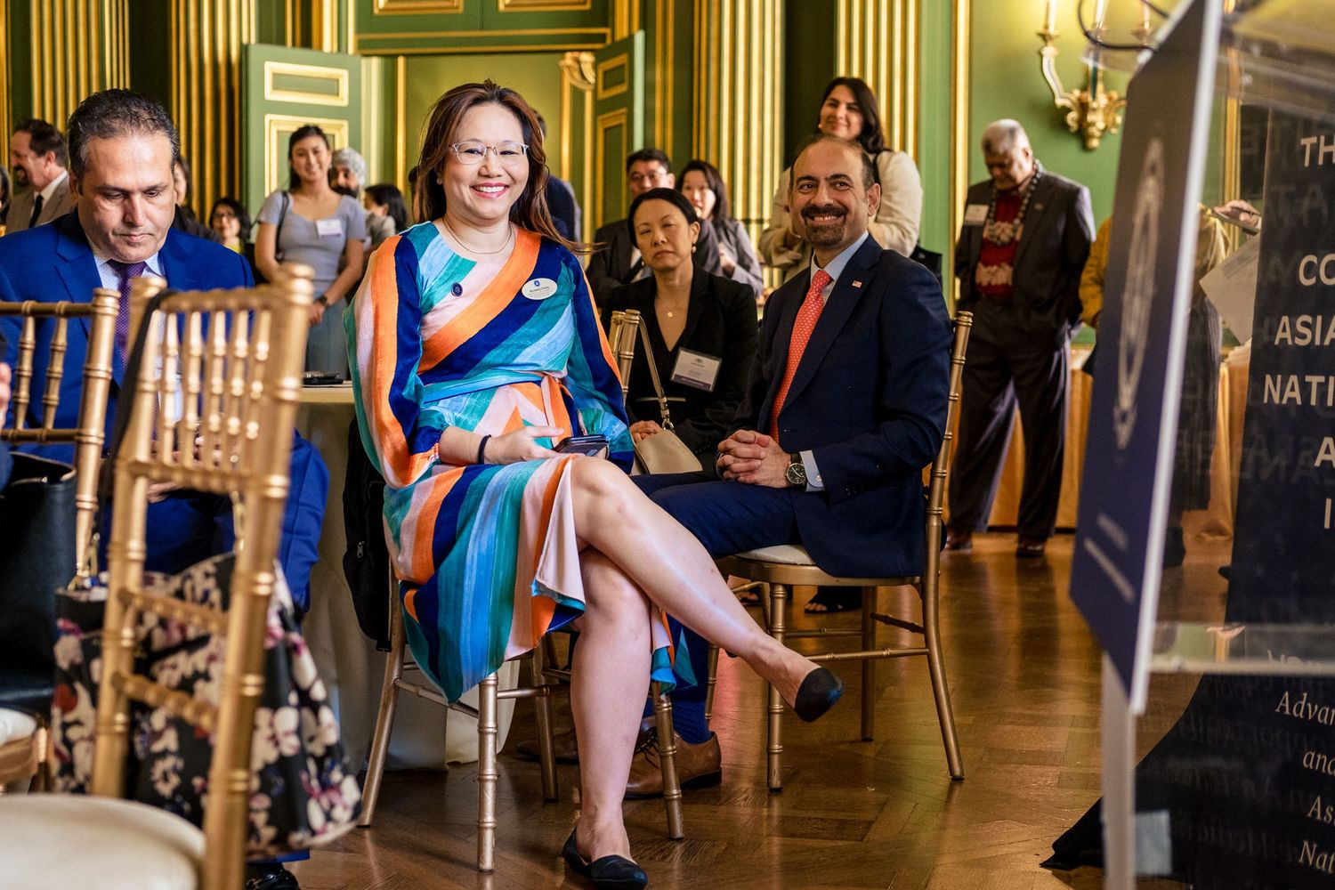 Woman in colorful dress seated at a formal event beside a man in a red tie in an ornate venue.