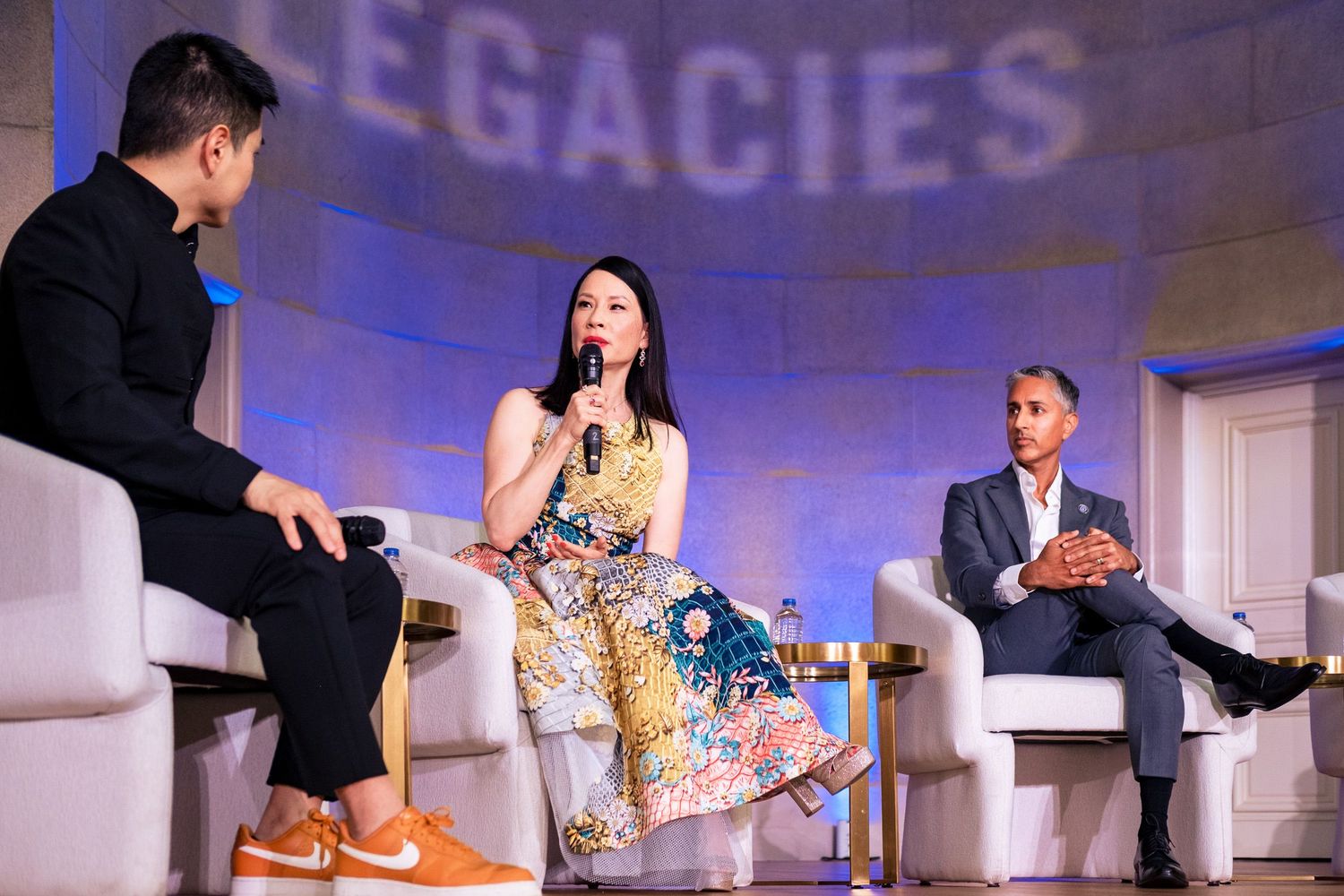 Three panelists on stage at a Legacies event, two seated and one speaking into a microphone.