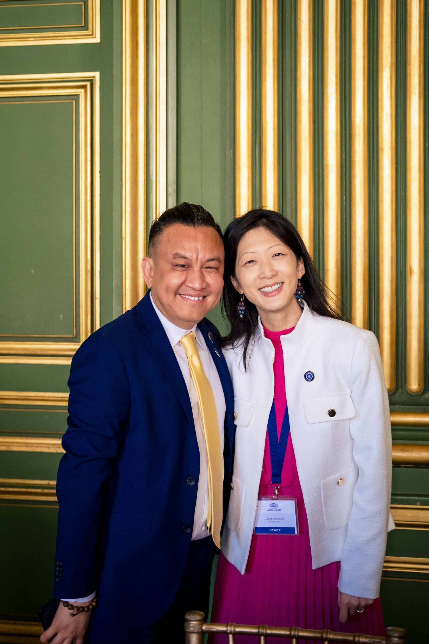 Two professionals smiling together in elegant green and gold room, man in blue suit, woman in white blazer with conference badge.