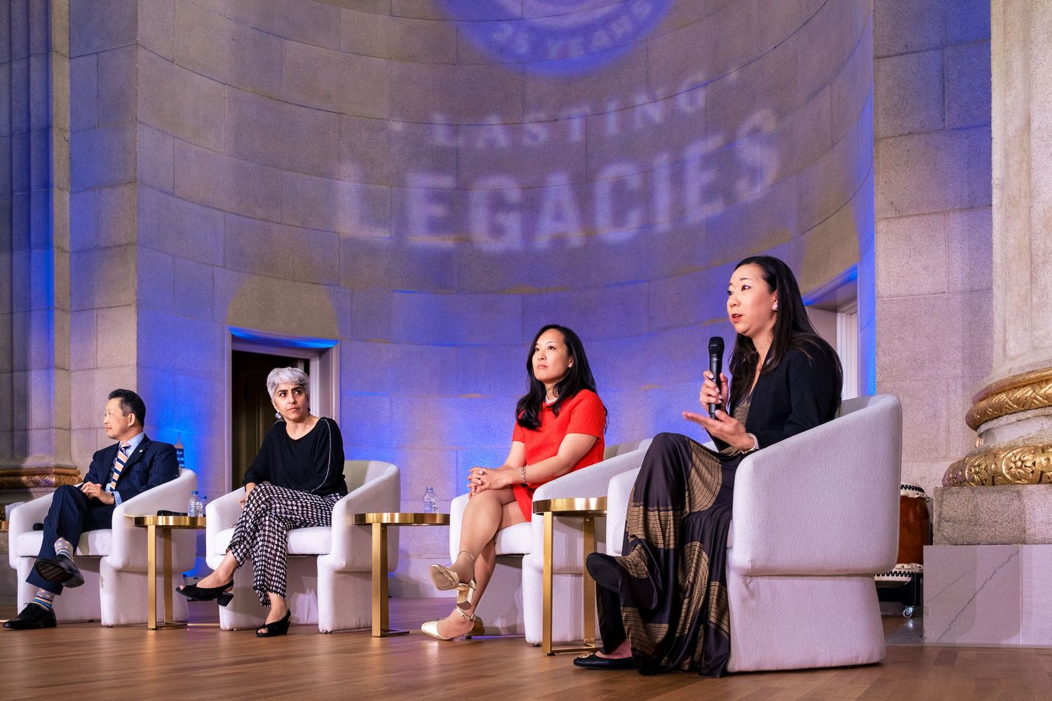Four panelists speak on stage at a Lasting Legacies event, seated in white chairs under elegant blue-lit decor.