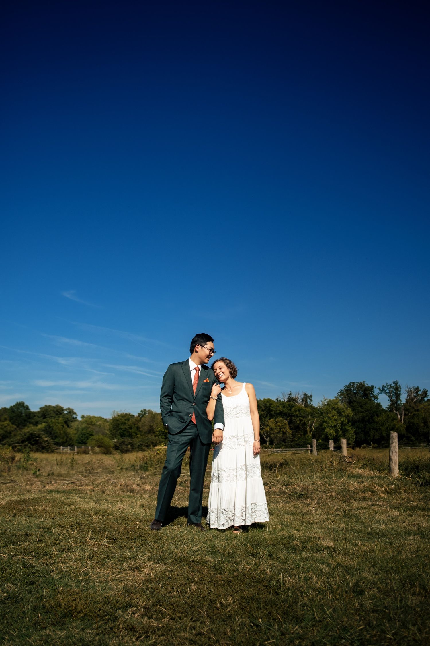 A newlywed couple stands together in a sunlit open field under a vivid blue sky.