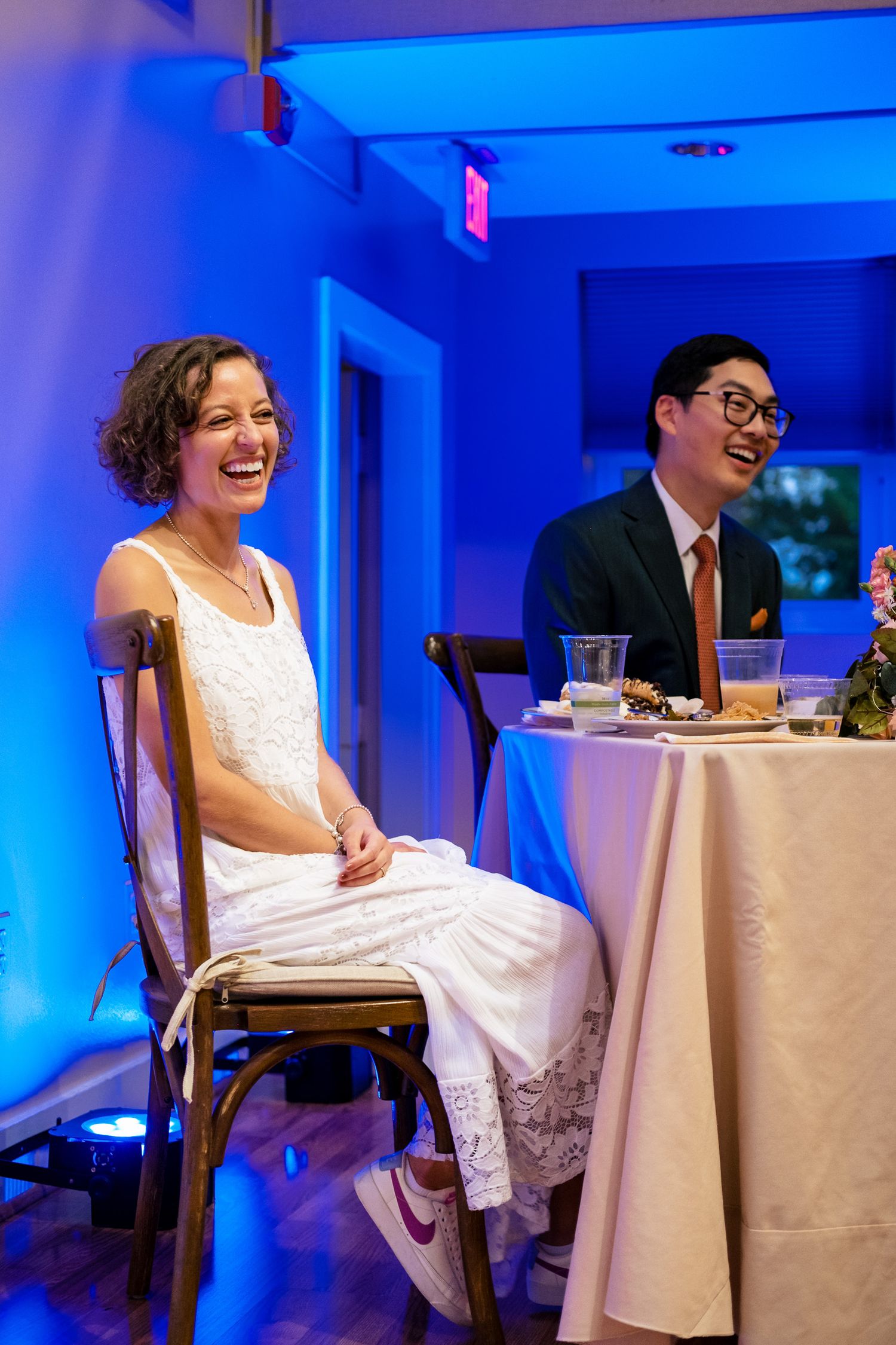 Bride in white dress and groom in navy suit laughing at wedding reception table.