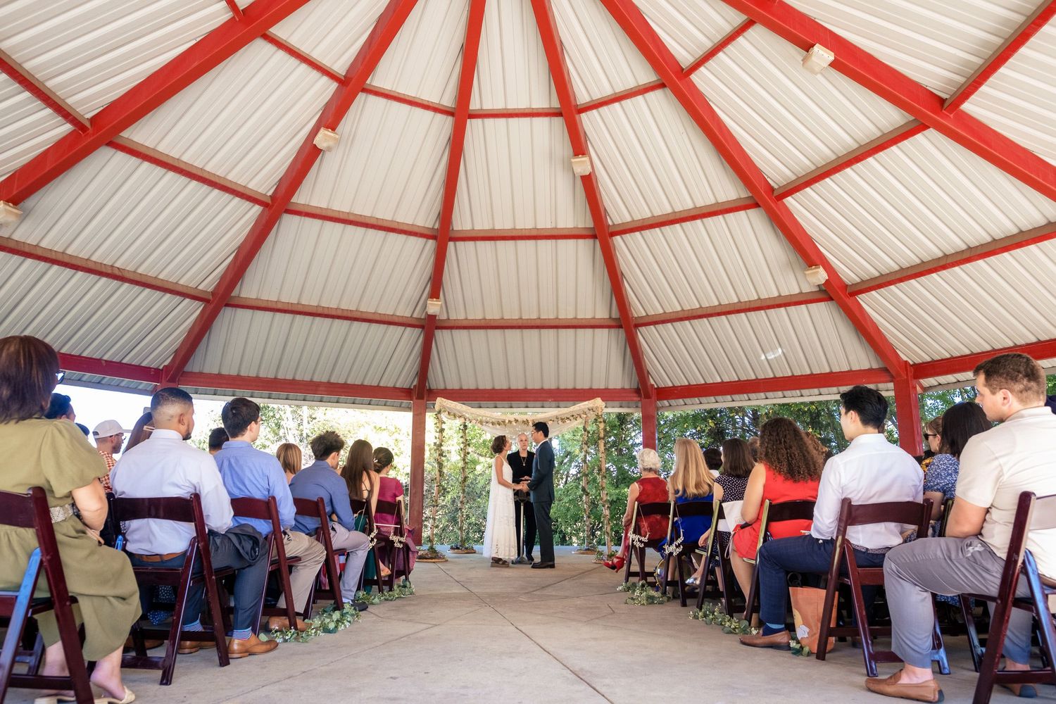 Wide angle view of wedding guests seated in a red and white gazebo during an outdoor ceremony.