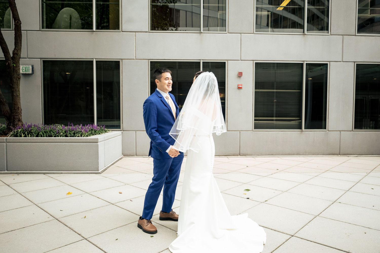 Groom in blue suit gazes at bride in flowing white wedding gown with long veil outdoors.
