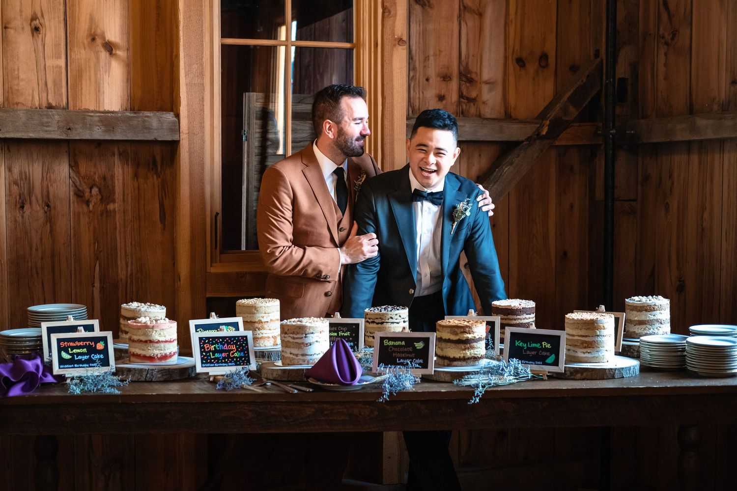 Two grooms laugh together at rustic barn wedding cake dessert table display.