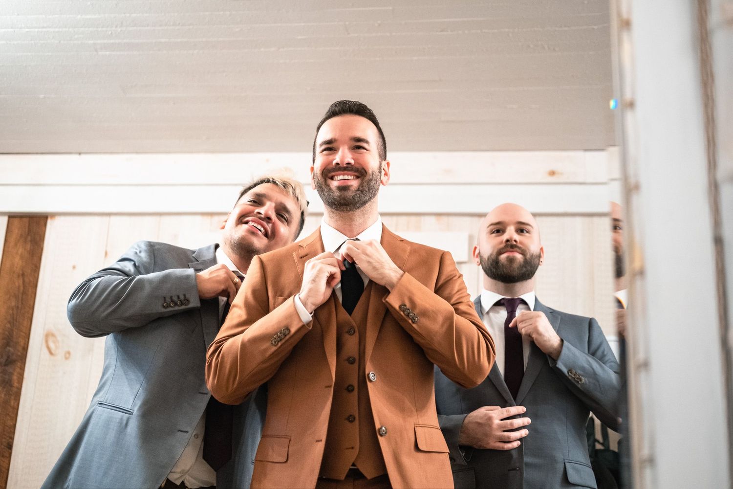 Groom in brown suit adjusting bow tie with two groomsmen in gray suits helping him get ready.