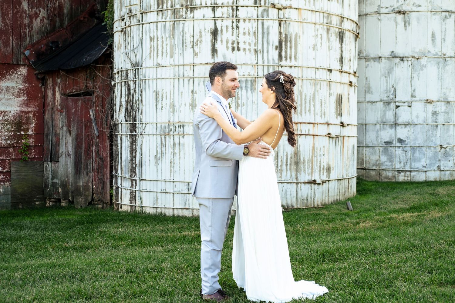 Bride and groom embrace tenderly while facing each other in front of rustic barn and silos.