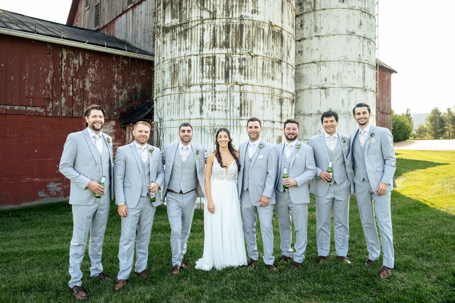 Bride in white gown poses with groomsmen in light gray suits beside a rustic barn silo.
