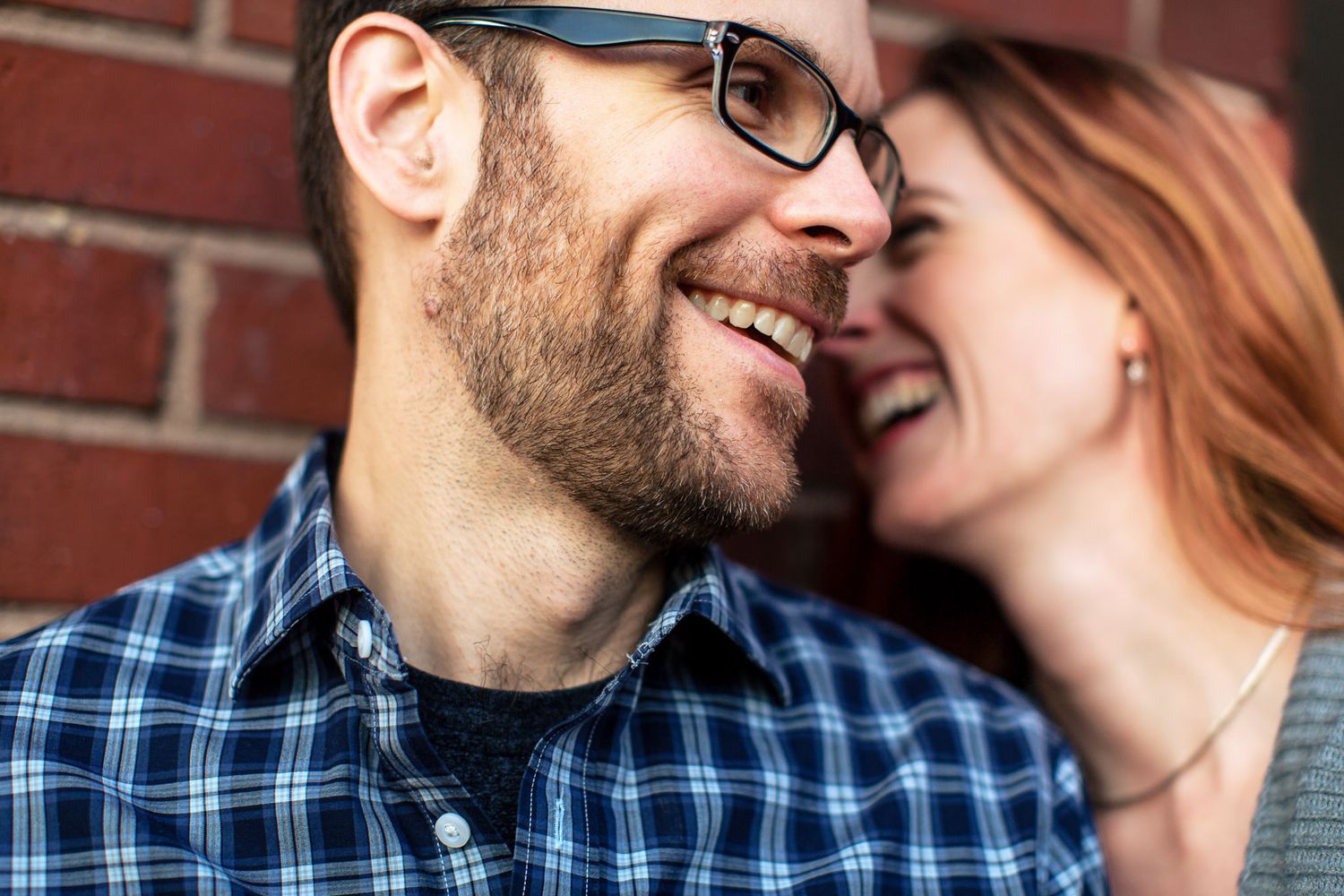 Smiling man with glasses shares a joyful moment with redhead woman by brick wall.