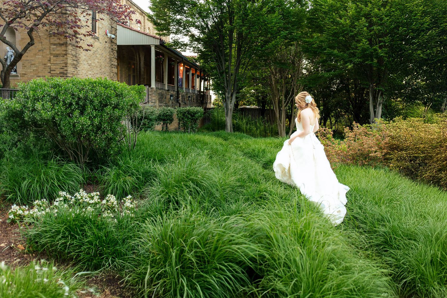 Bride in flowing white gown walks through lush green garden landscape beside a historic building.