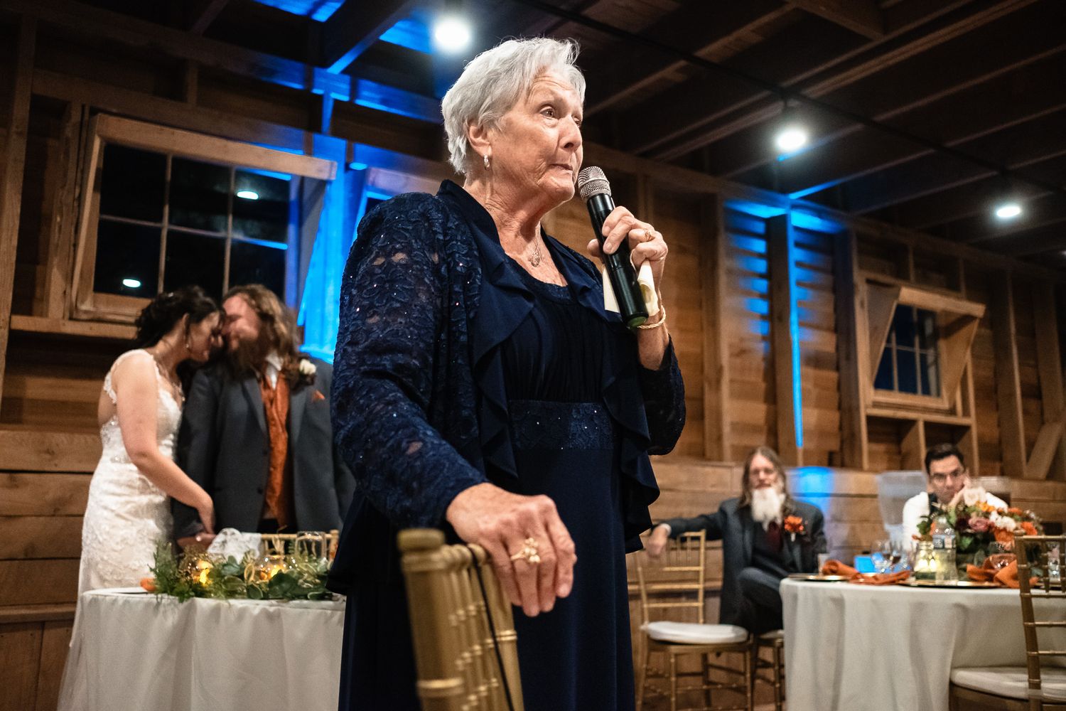 Elderly woman in navy blue dress gives a heartfelt speech at a rustic barn wedding reception.