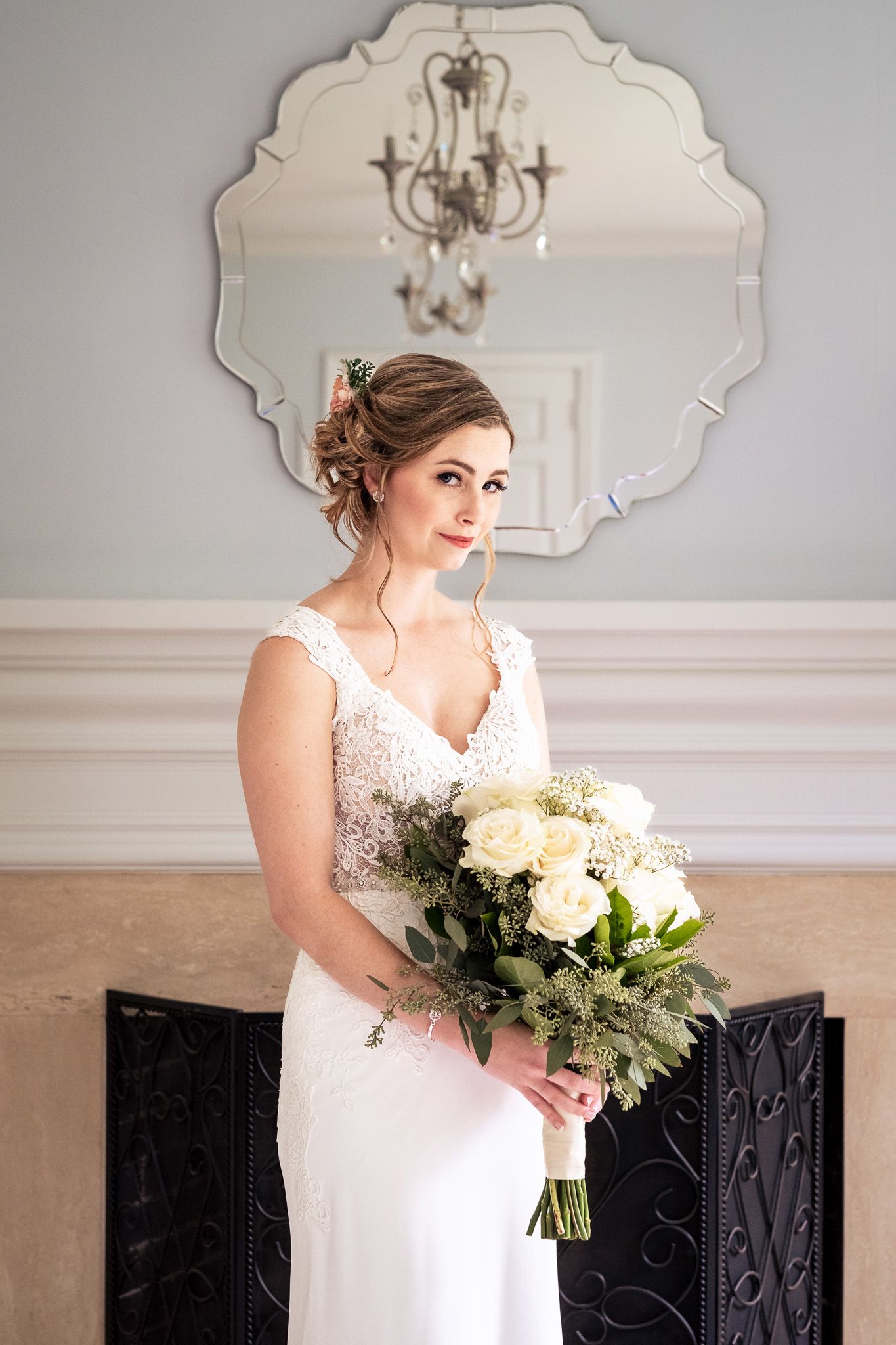 Bride in white lace gown holds bouquet of white roses in front of ornate mirror.