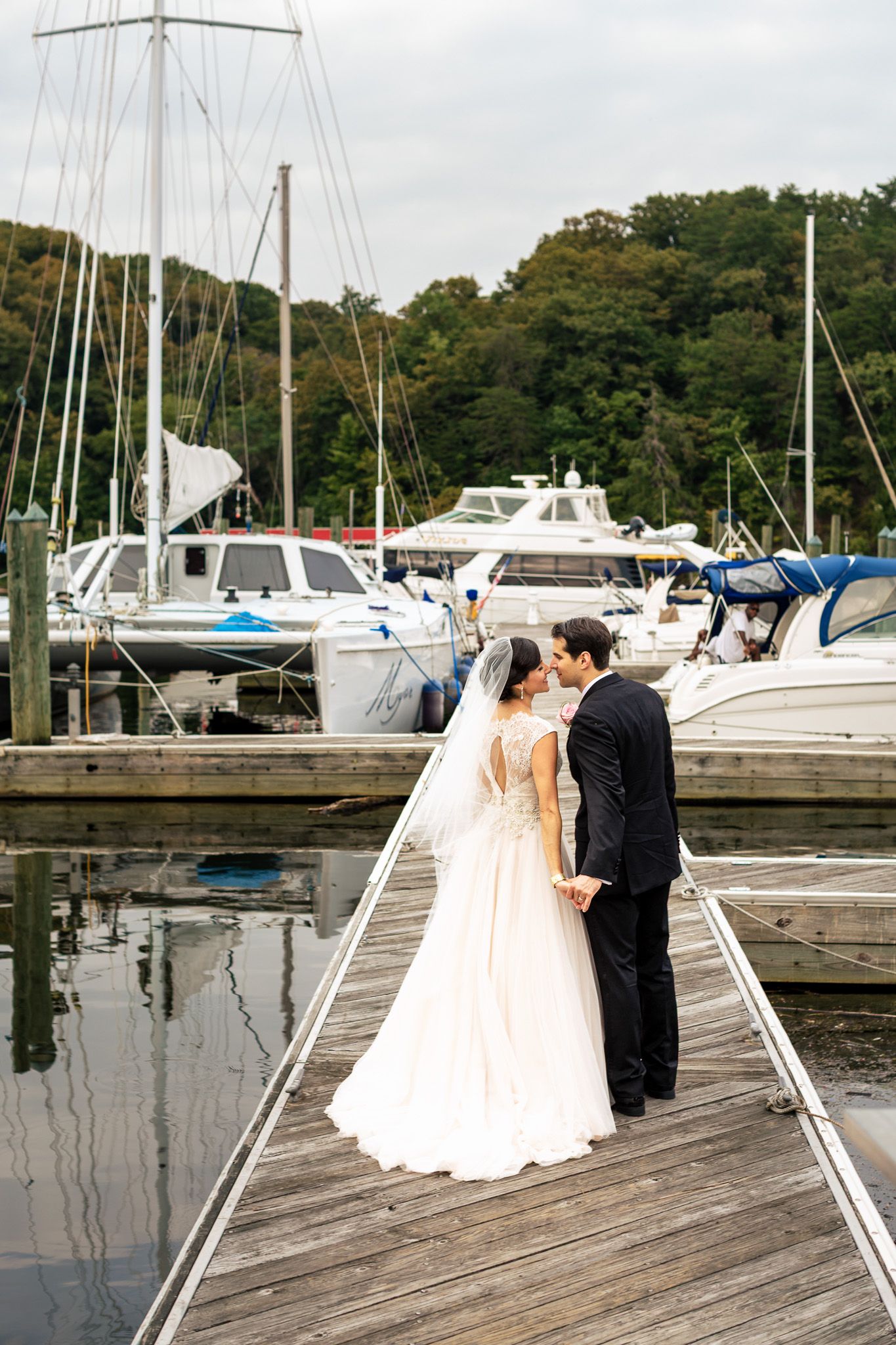 Bride and groom share a romantic moment on a wooden dock surrounded by sailboats at marina.