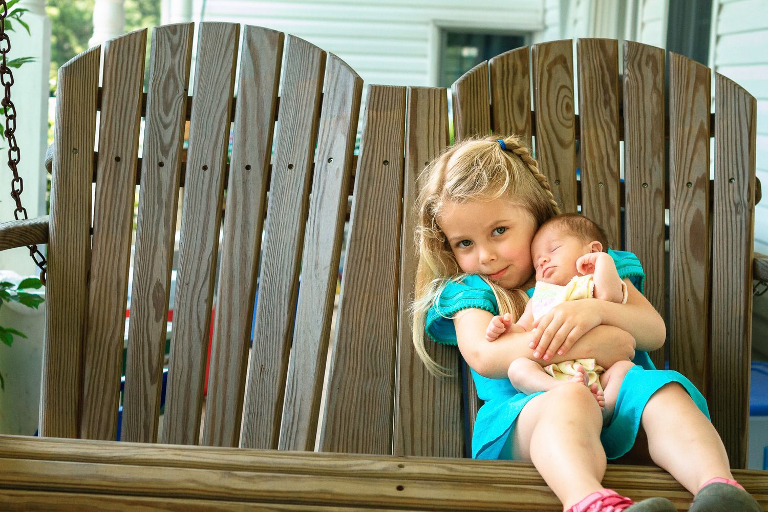 Young blonde girl sitting on a wooden porch swing gently cradling a sleeping newborn baby in her arms.