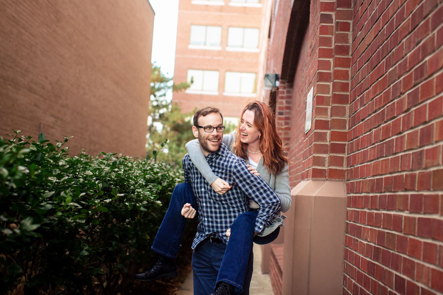 Man giving woman a piggyback ride while laughing near a brick building outdoors.
