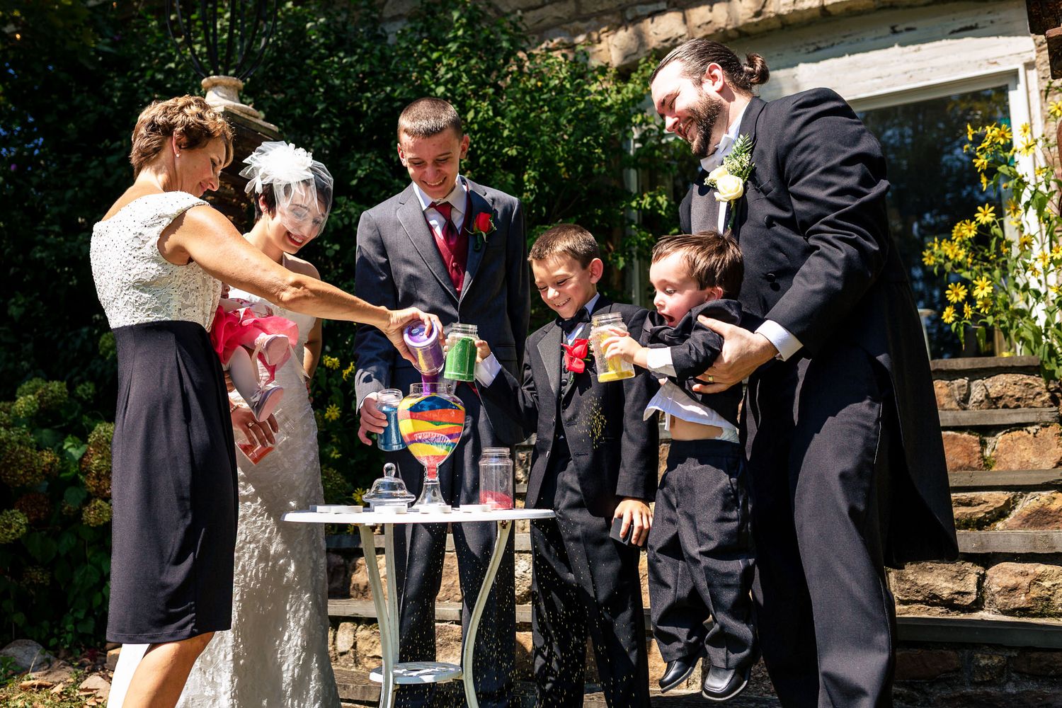 Family performing colorful sand ceremony at outdoor wedding with children in formal attire.