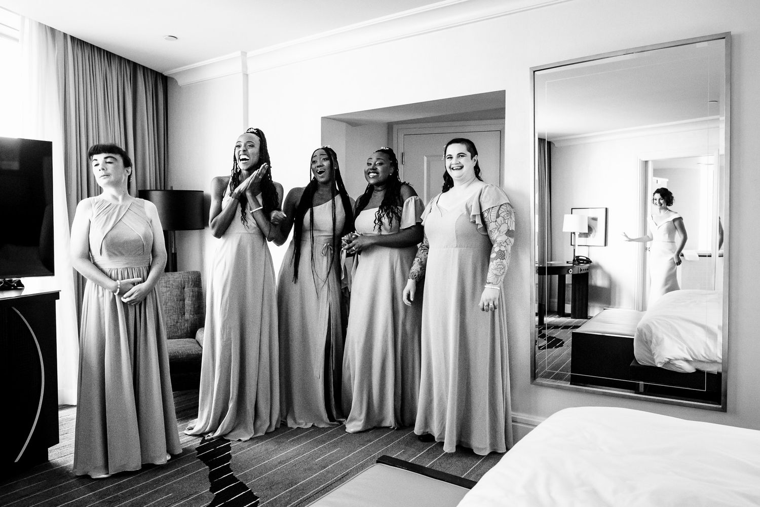Four bridesmaids in gray gowns react with joy as bride prepares in hotel room.