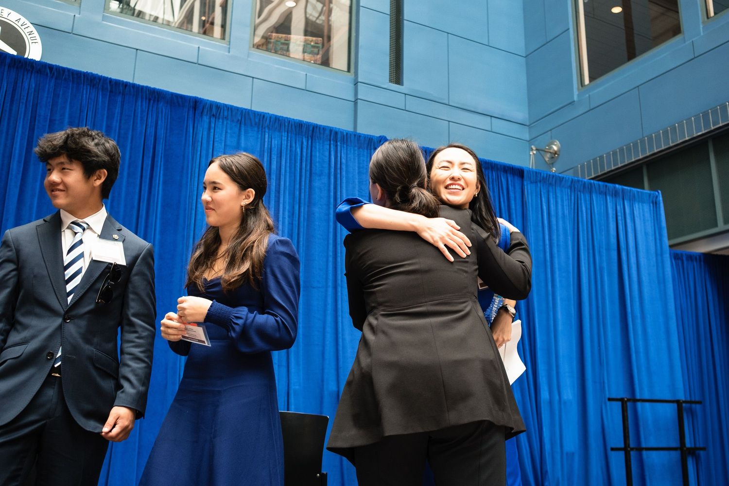 People hugging and smiling on stage at a formal event with blue curtain backdrop.