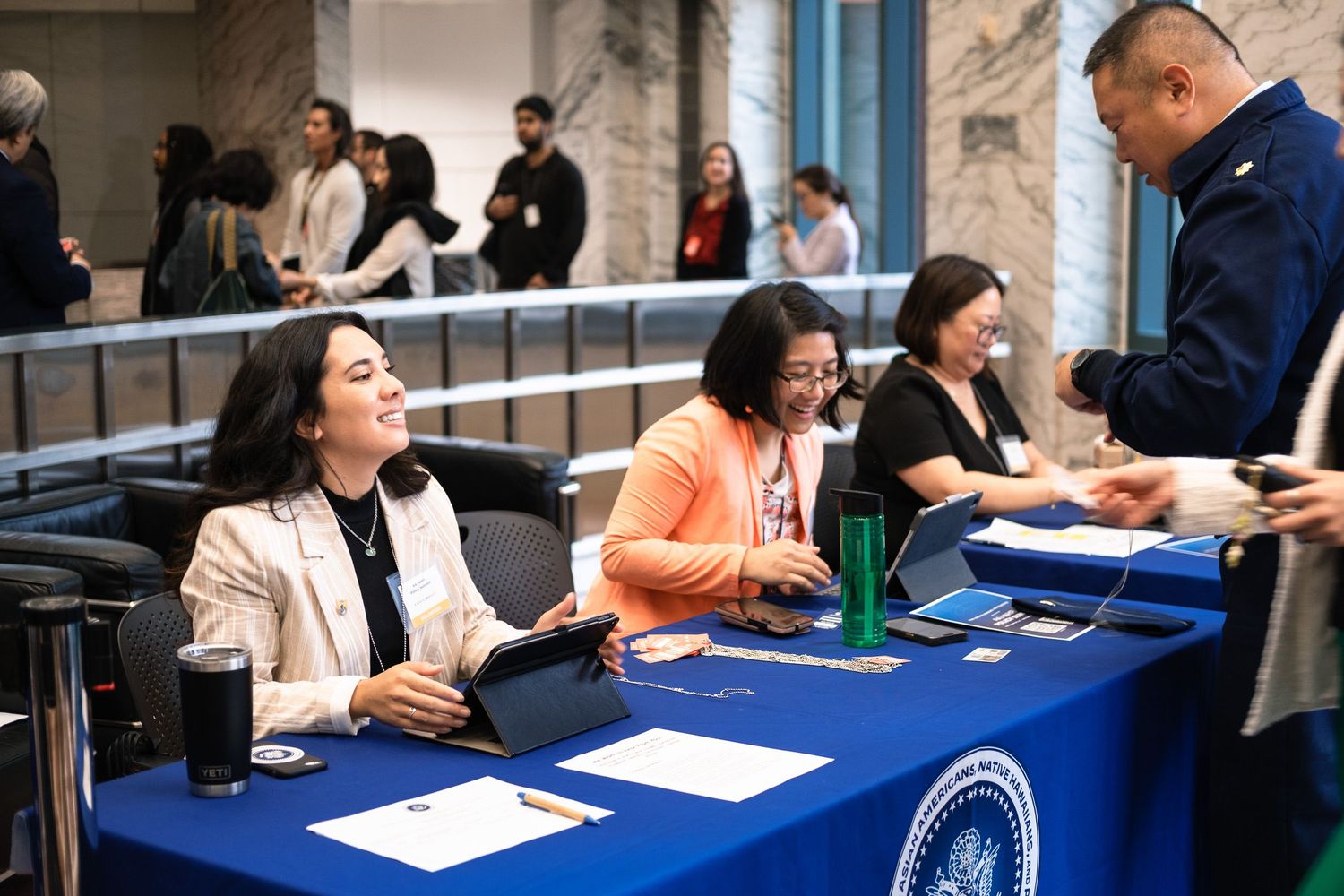 Women smile at a registration table with a blue tablecloth at a professional conference event.