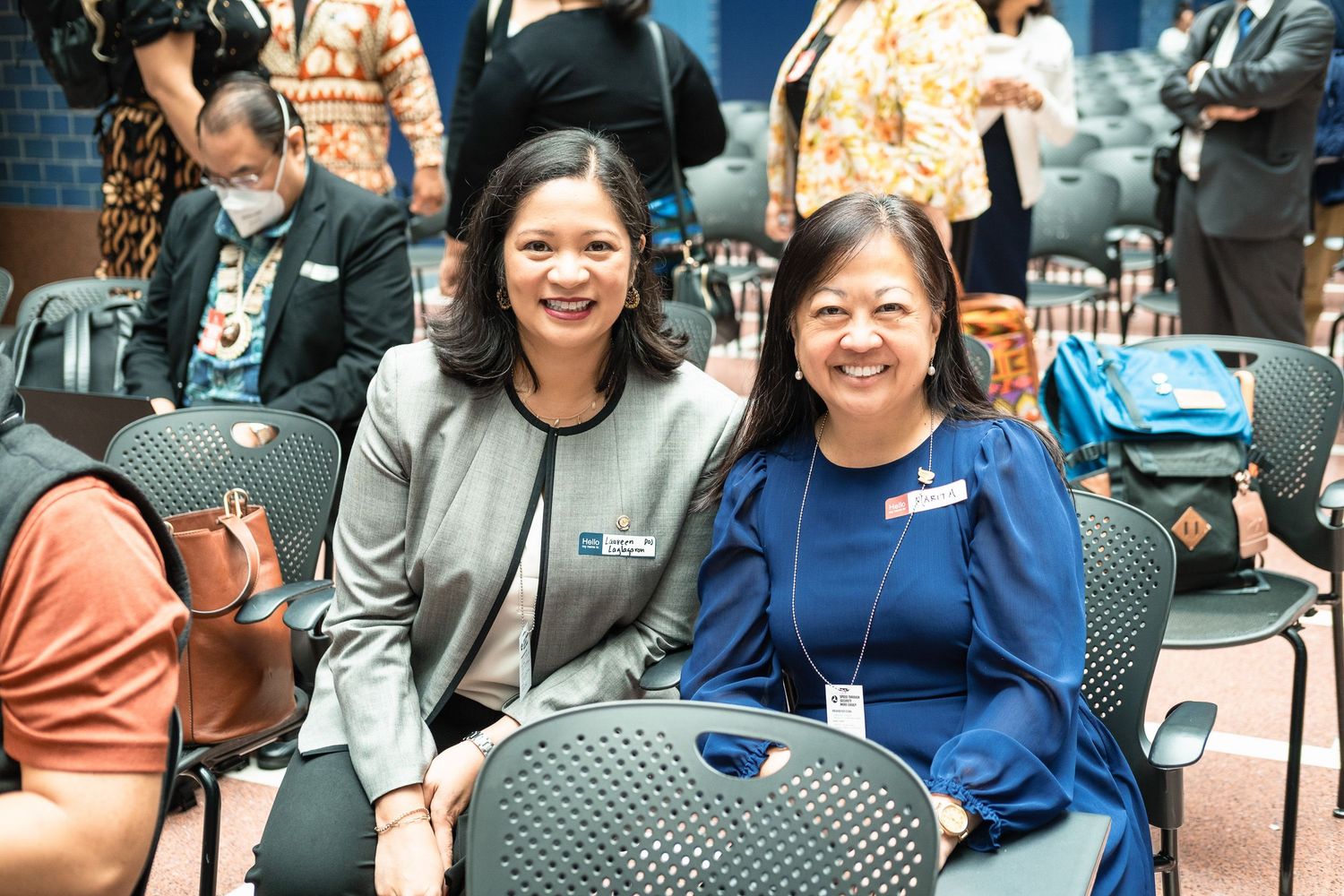 Two smiling women pose together at a professional event, seated in chairs at what appears to be a conference or meeting.