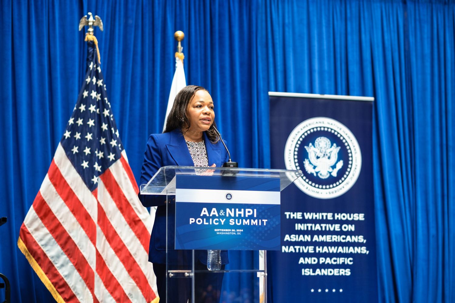 Woman in blue suit speaks at AA&NHPI Policy Summit podium beside American flag and White House Initiative sign.