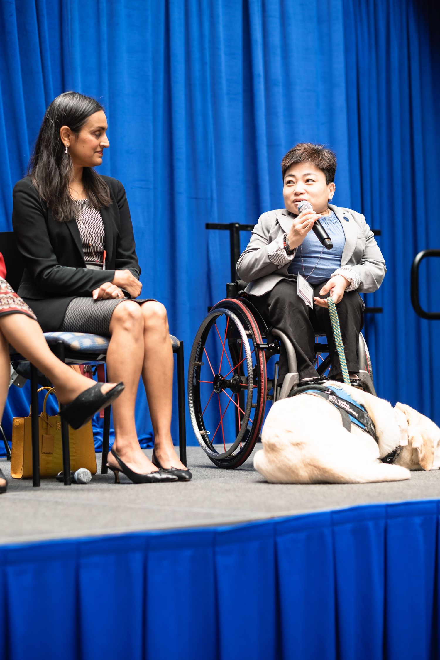 Two women on stage, one in a wheelchair speaking into a mic with a service dog nearby, at a professional event.
