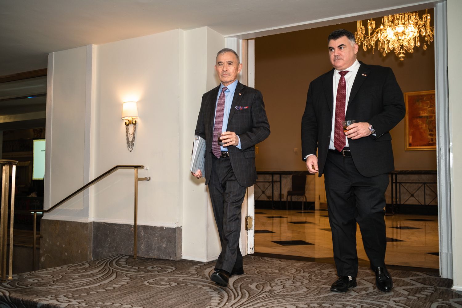 Two businessmen in suits walking through an elegant hotel corridor with chandelier lighting.