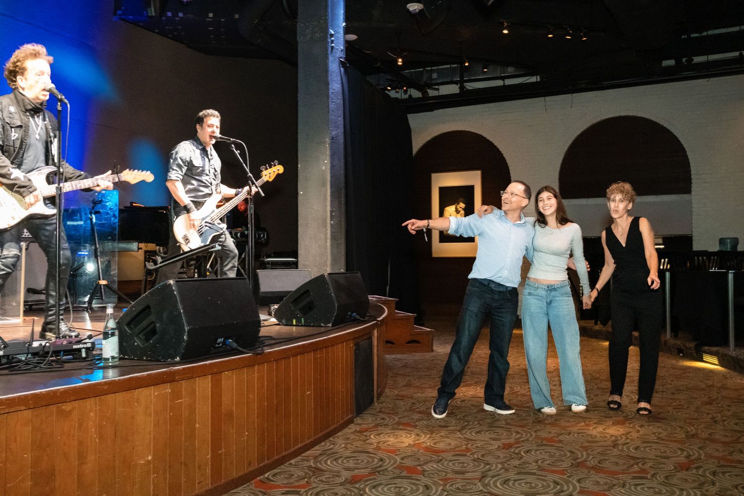 Live band performs on stage while three guests pose and point in a stylish indoor venue with patterned carpet.