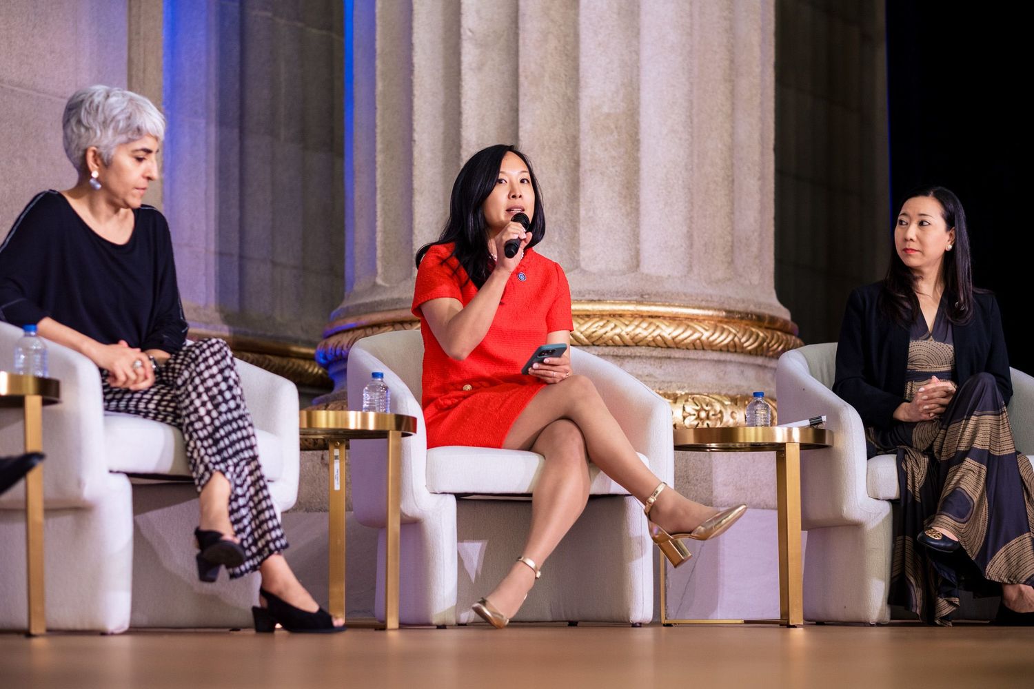 Three women participate in a panel discussion at an elegant venue, with one in red speaking into a microphone.