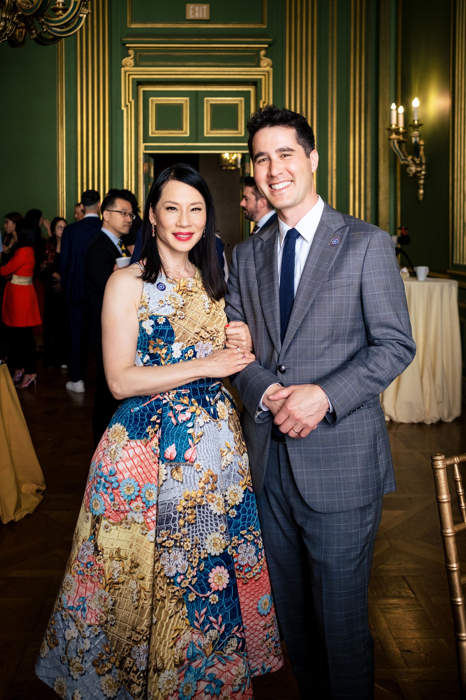 A woman in a colorful floral dress and a man in a gray suit smile together at an elegant green-walled event venue.