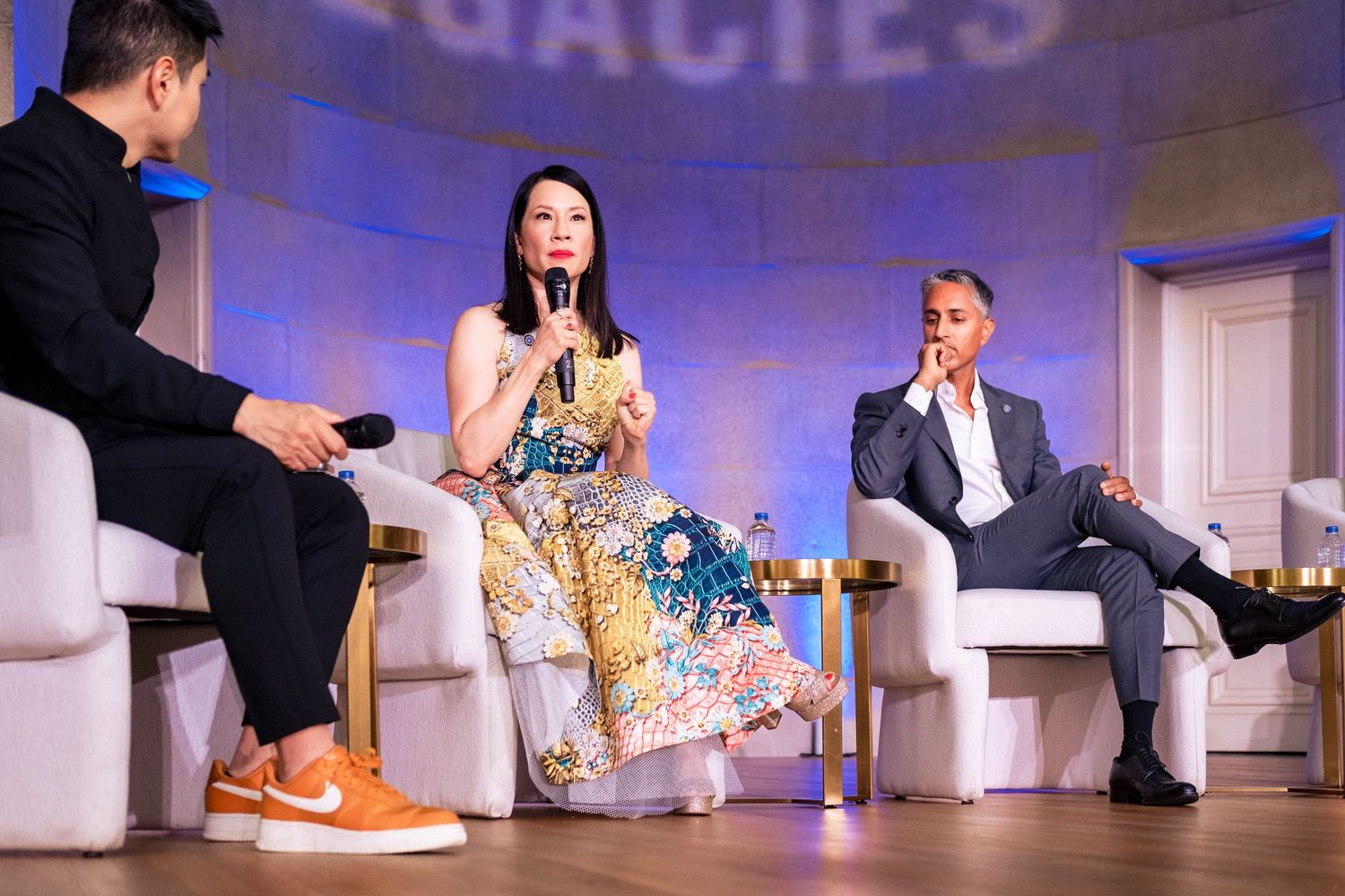 Three panelists engaged in discussion on stage, woman in floral dress speaking into microphone at elegant event.