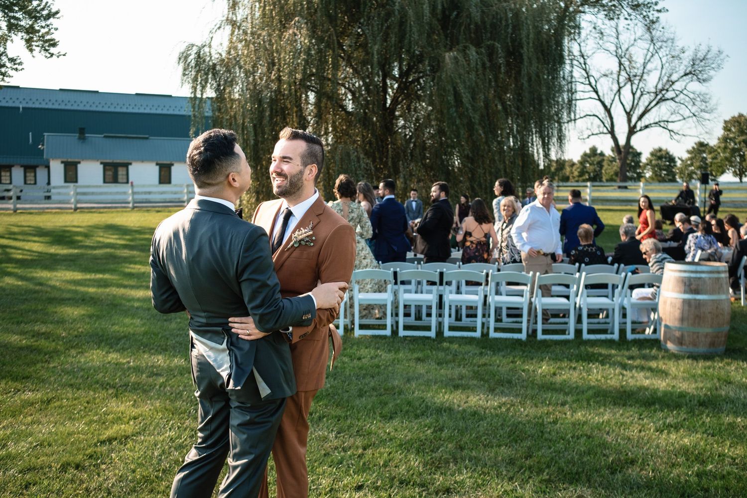 Two grooms embrace and smile together after their outdoor wedding ceremony near a rustic barn venue.