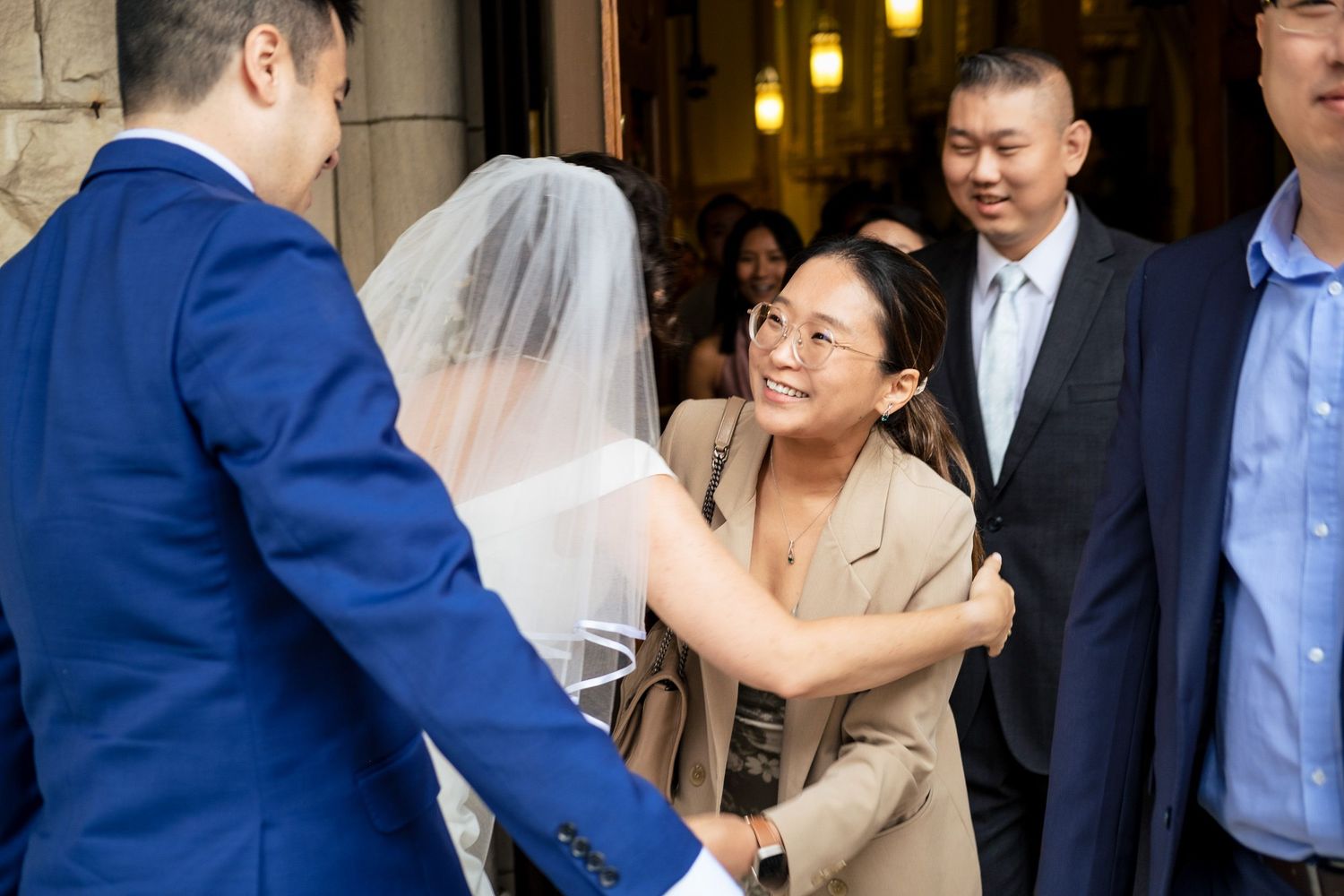 Wedding guest in beige jacket warmly greeting groom in blue suit as bride in veil looks on at church steps.