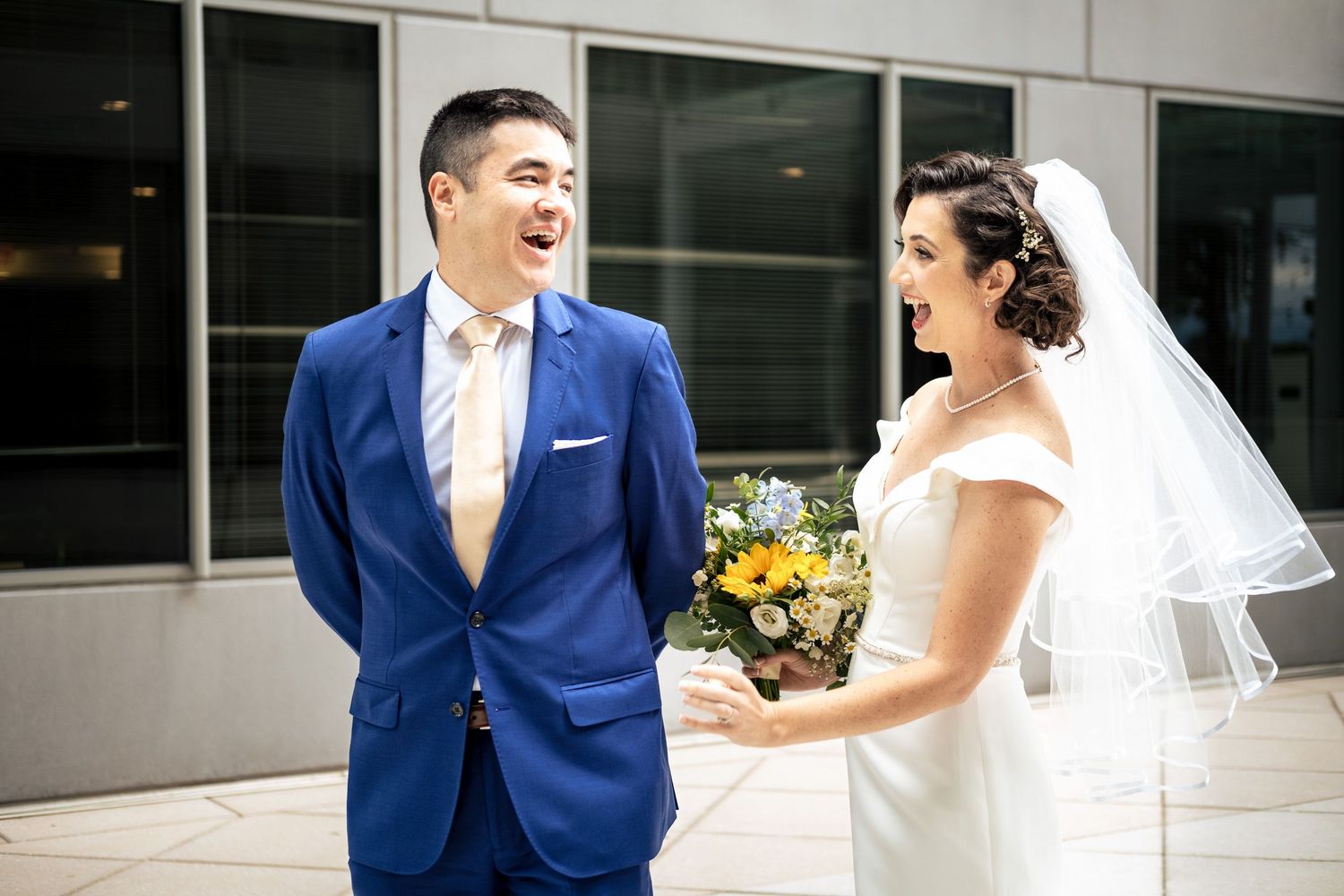 Bride and groom share a joyful laugh during first look, bride holding colorful bouquet in urban setting.