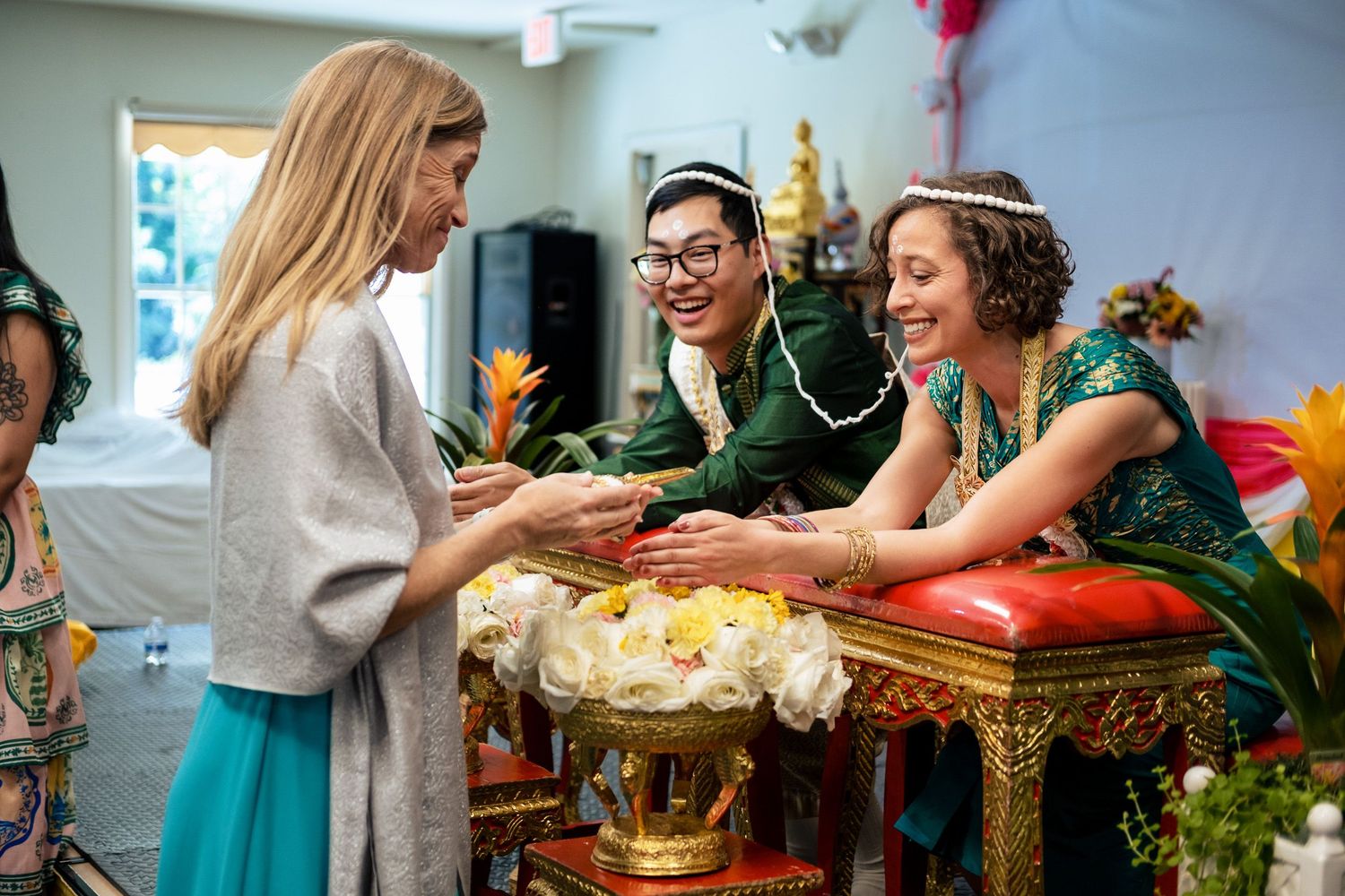 Guest presenting flowers to smiling couple in traditional Thai wedding attire during blessing ritual.