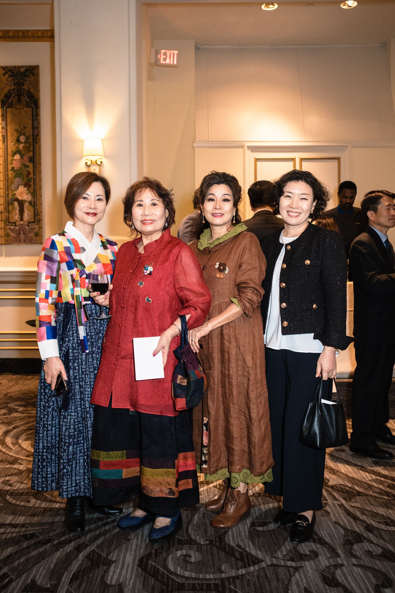 Four elegantly dressed women posing together at a formal indoor event, wearing colorful traditional and modern outfits.