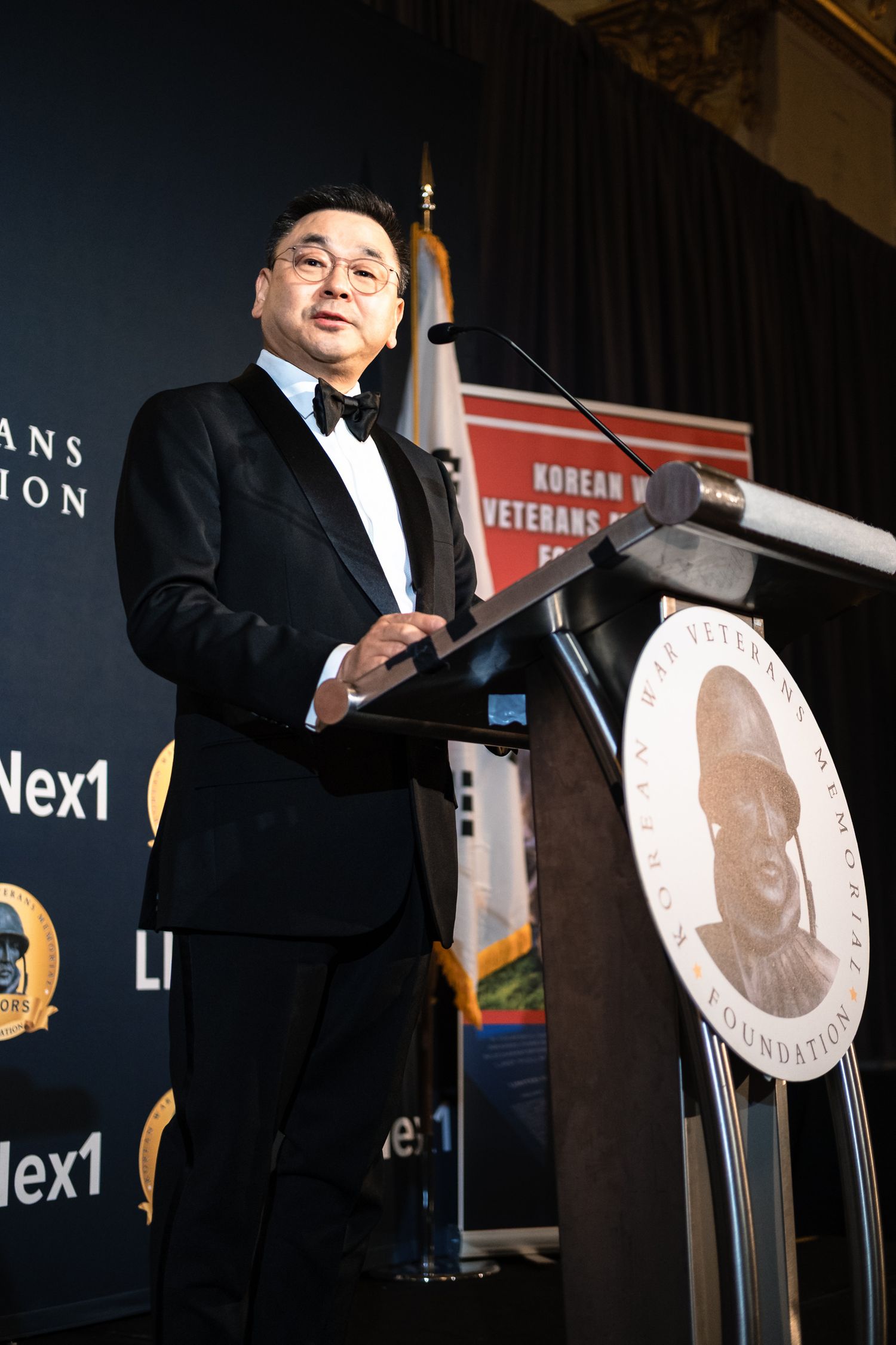 Asian man in tuxedo delivers speech at Korean War Veterans Memorial Foundation podium during gala.