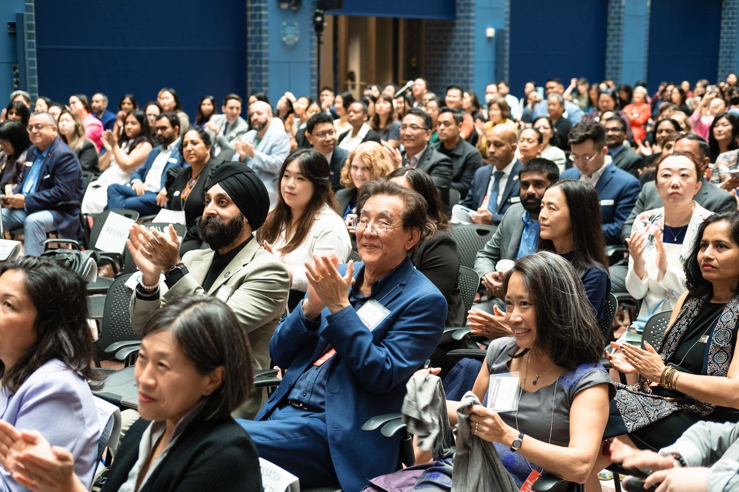 Diverse audience applauding at a conference event in a packed auditorium with attendees of various backgrounds.