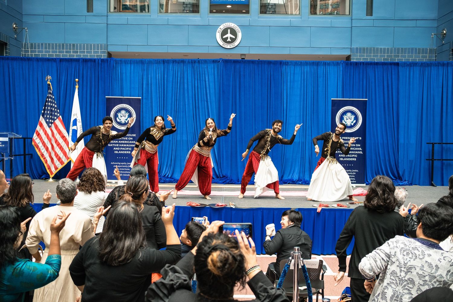 Indian classical dancers perform on stage before an audience at a formal event with US and organizational flags.