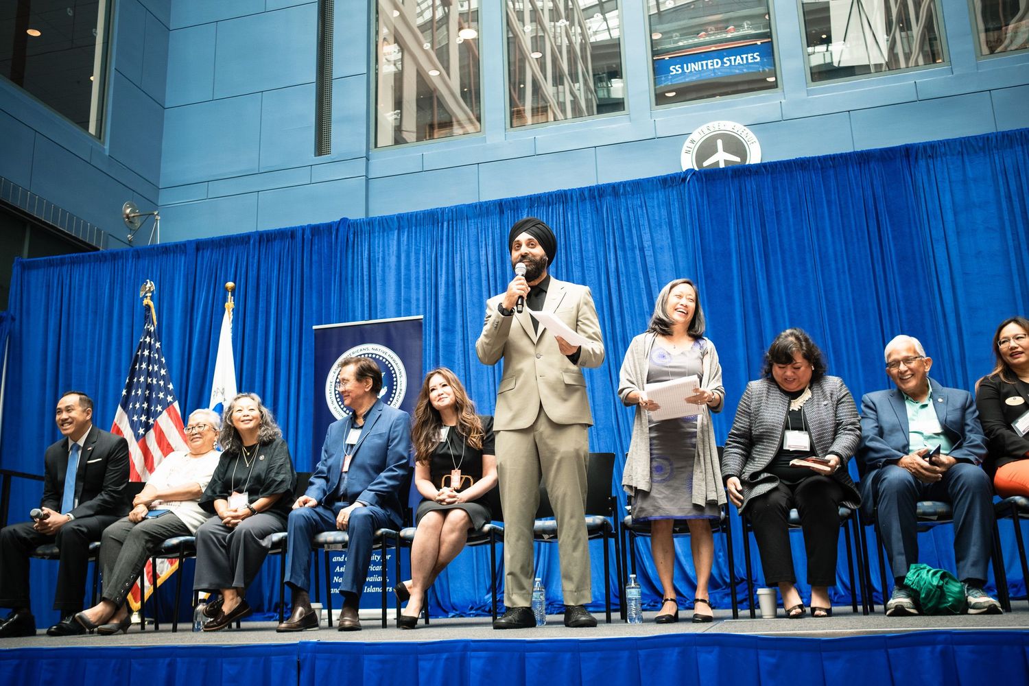 Man in tan suit speaks at podium on stage with seated panelists before blue curtain backdrop at formal event.