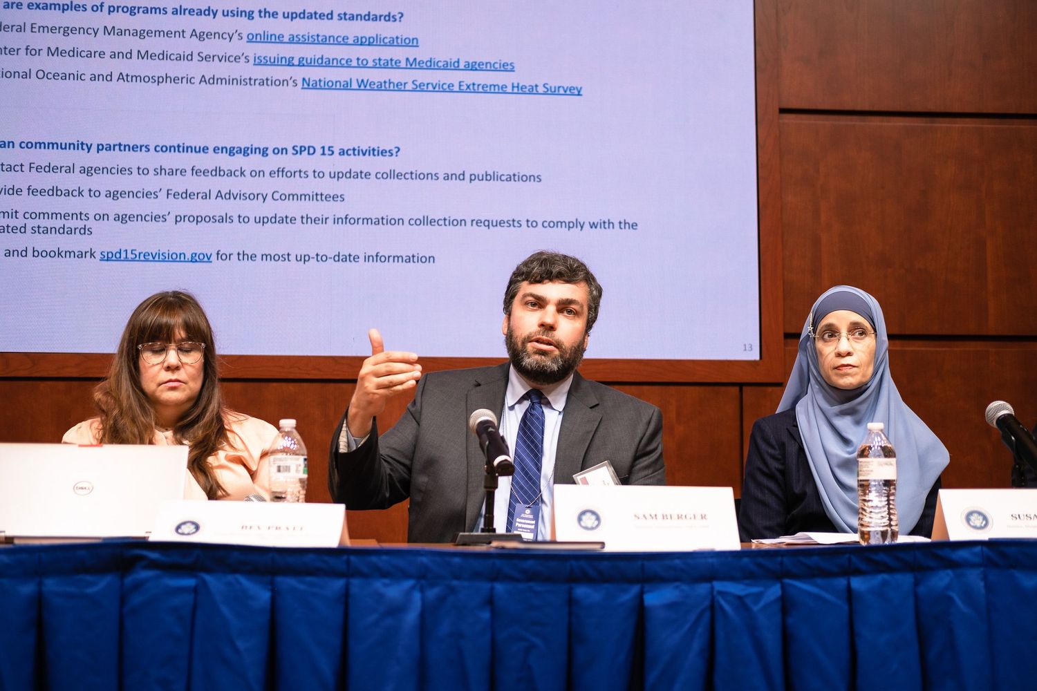 Three panelists at a conference table, man in center speaking, women on either side, presentation screen visible behind them.