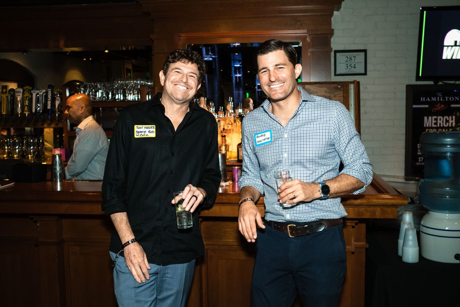 Two men smiling and holding drinks at a bar during a networking event.