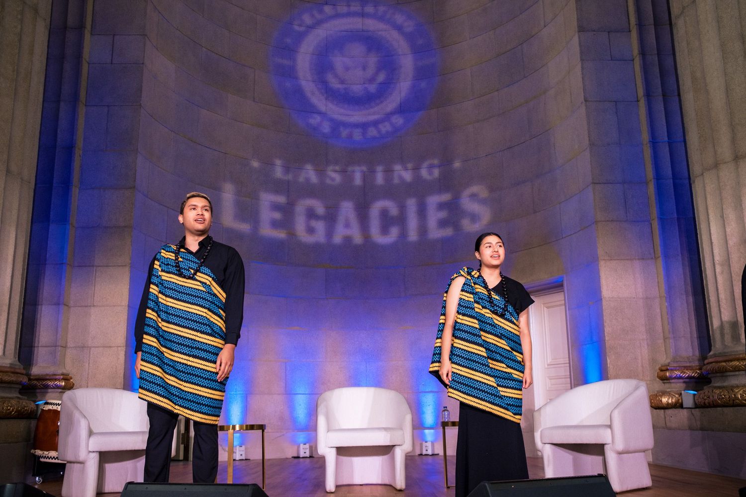 Two people in colorful striped robes stand on stage beneath a Lasting Legacies projection at a formal ceremony.
