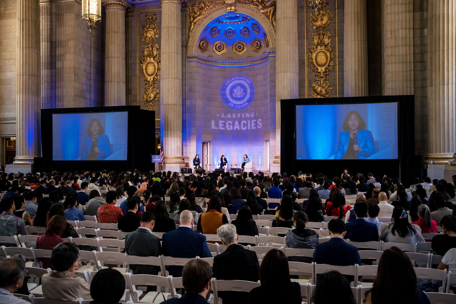 Large audience seated in ornate hall watching speaker on dual screens at Lasting Legacies event.